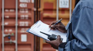 A worker holds documents on a clipboard and a walkie talkie in front of shipping containers