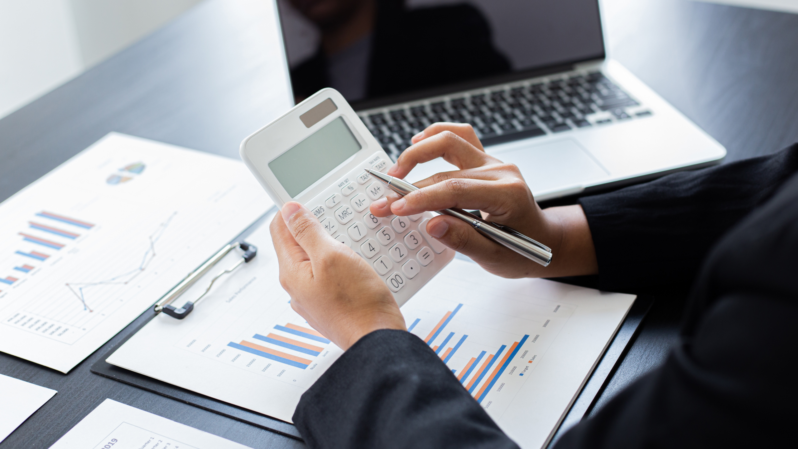 A person uses a calculator and a laptop atop several documents with charts and figures